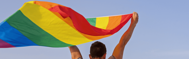 Foto de um homem branco com cabelo curto e braços levantados segurando a bandeira do movimento LGBTQIA+ #MêsDoOrgulhoAutista #MêsDoOrgulhoLGBTQIA #8M